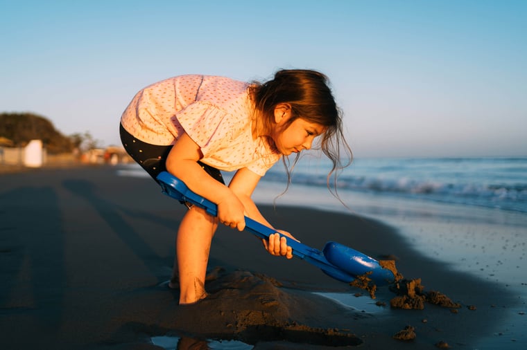 A young child digs in the sand with a blue shovel on the beach, with the evening light casting a soft glow on the activity.